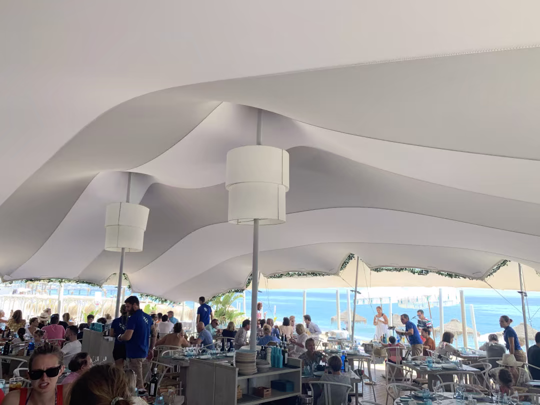 Busy hospitality area with guests seated beneath a white stretch tent beside the ocean